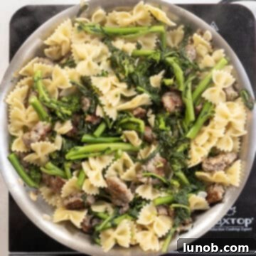 Al dente farfalle pasta tossed into the pan with sausage, broccoli rabe, and sauce, simmering together to finish the dish.