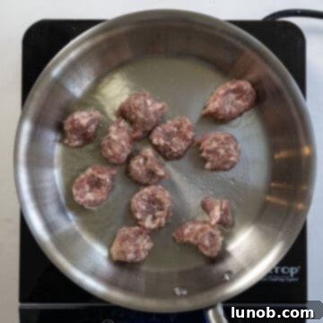 Pieces of sweet Italian sausage searing in a hot pan with olive oil, developing a golden-brown crust.