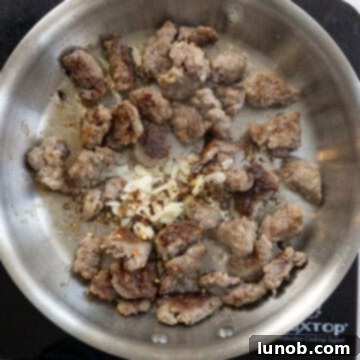 Sautéed Italian sausage mixed with minced garlic and crushed red pepper flakes in a pan, forming the aromatic base of the sauce.