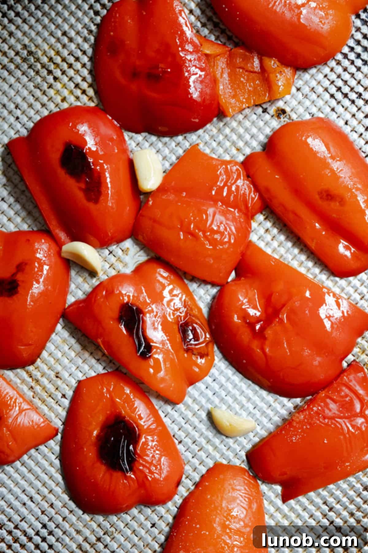 Fresh red peppers and whole garlic cloves roasting on a baking sheet in the oven.