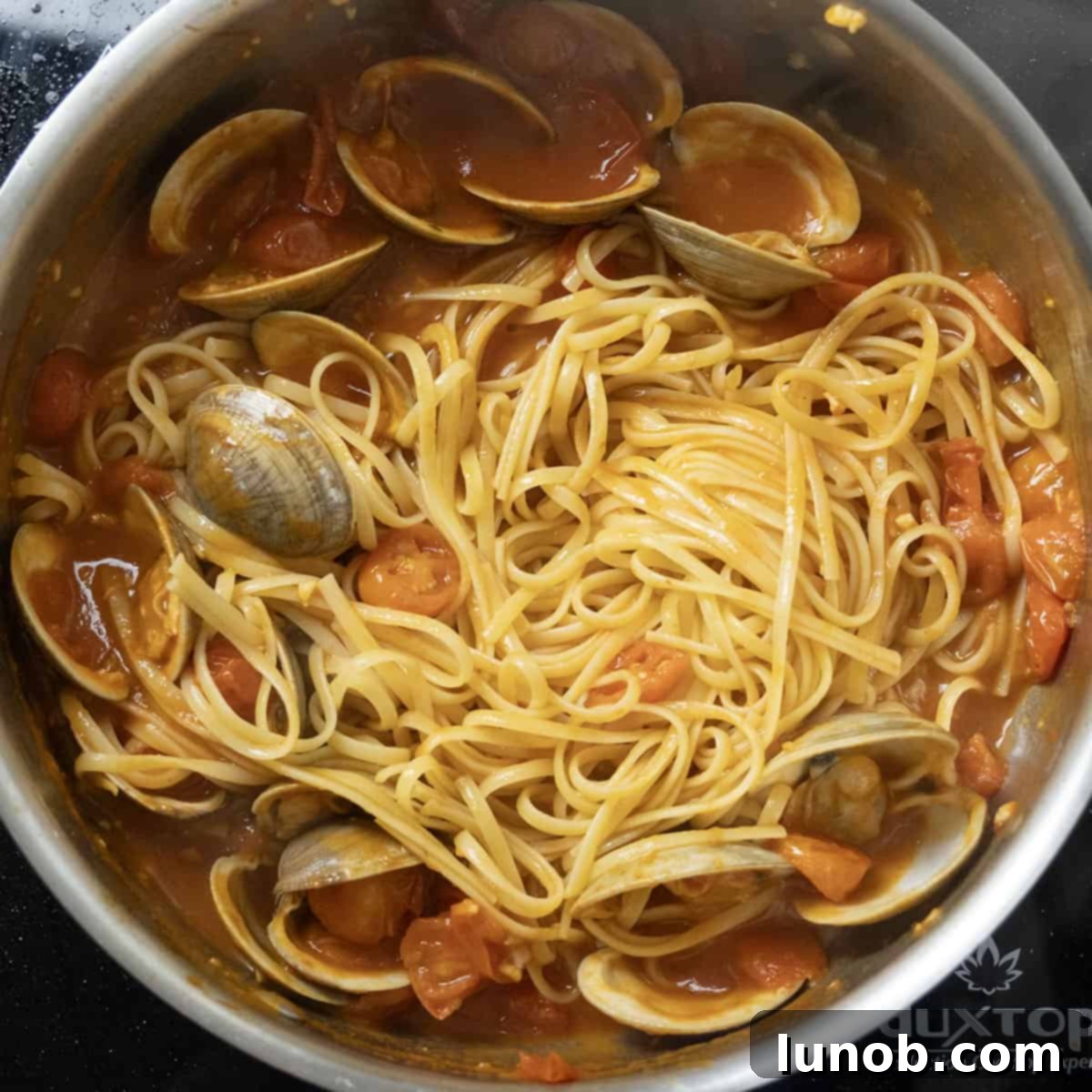 Cooked linguine being tossed into the red clam sauce in a large pan.