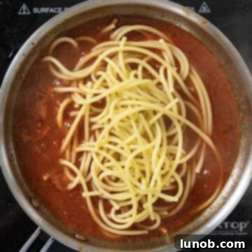 Partially cooked bucatini pasta being added to the red Amatriciana sauce in a saucepan.