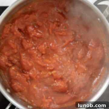 Whole peeled plum tomatoes being gently broken apart in a saucepan, mixed with basil and seasonings, simmering into a rich sauce.