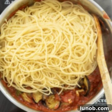 Cooked spaghetti being tossed into the rich tomato and eggplant sauce in a large saucepan.