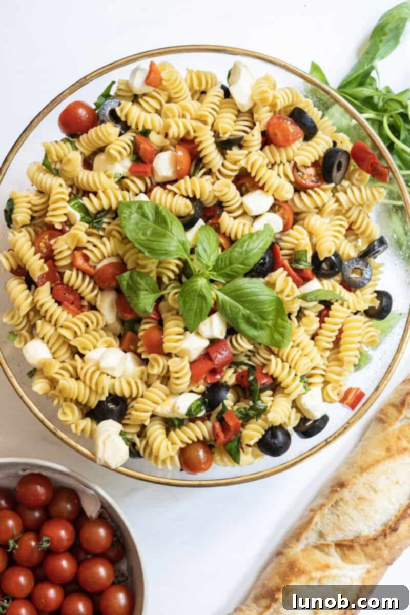 Italian pasta salad in a large bowl with garnish of basil, on the side is a loaf of bread and bowl of cherry tomatoes. 
