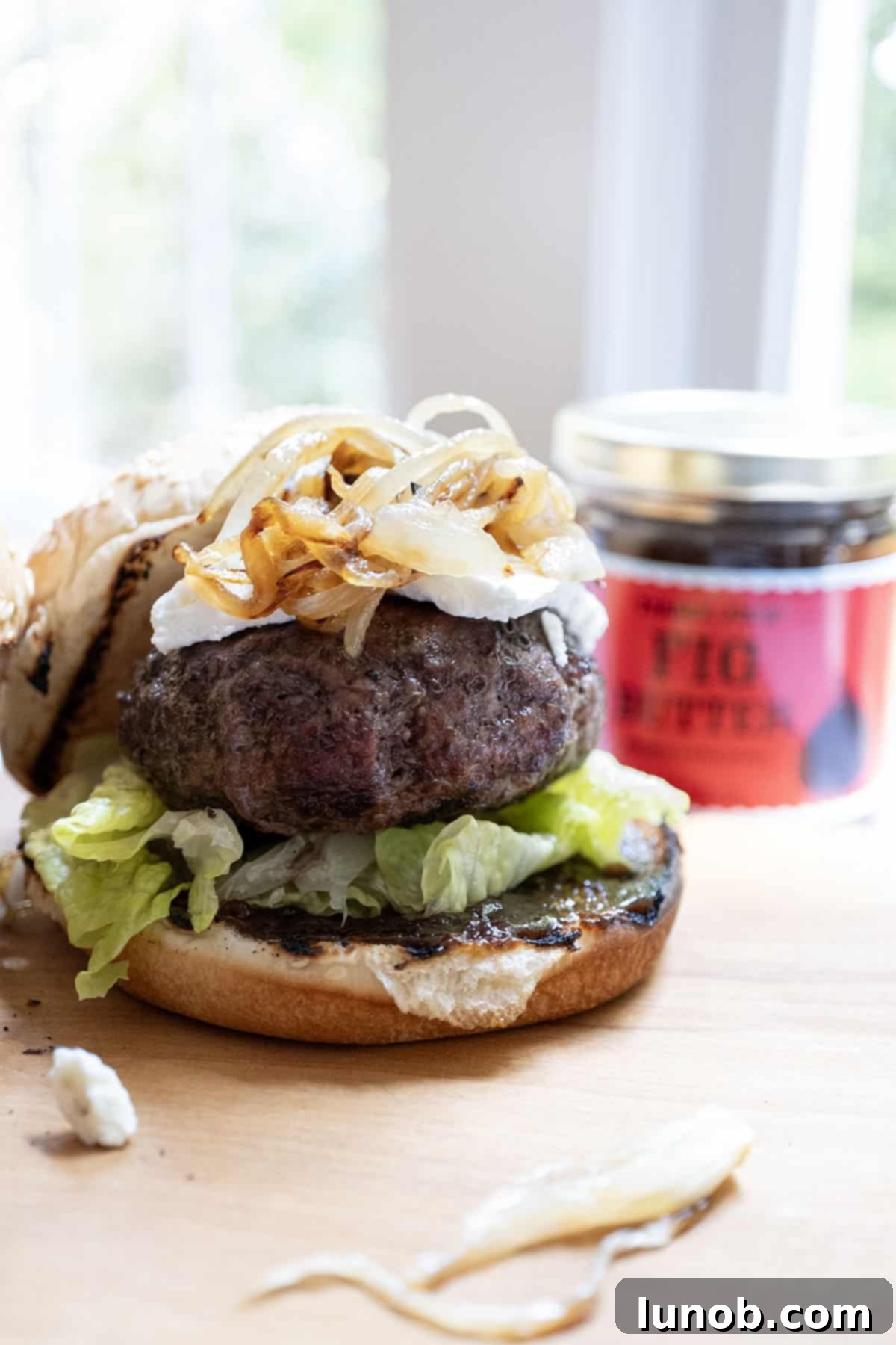 A gourmet burger featuring a juicy patty, a generous layer of sweet fig jam, creamy goat cheese, and golden caramelized onions, all nestled on a bed of fresh lettuce.