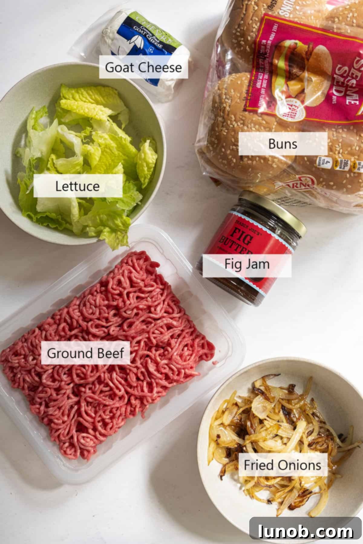 A curated selection of fresh ingredients laid out, including ground beef, goat cheese, fig jam, an onion, olive oil, butter, and fresh arugula, ready for the recipe.