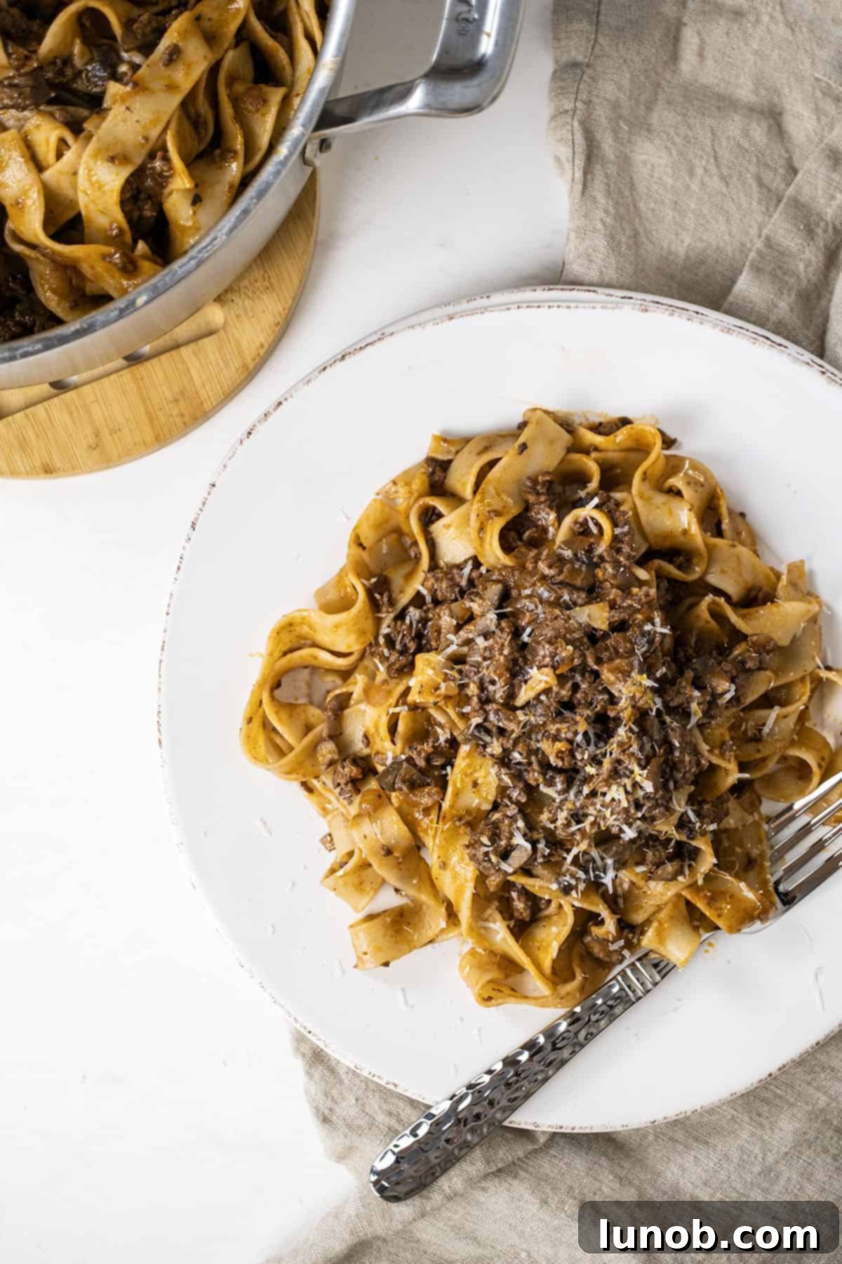 Close-up of lamb ragu with pappardelle on a white plate, with a large pan of pasta in the background.