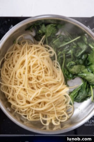 Cooked spaghetti being added to a saucepan with wilted spinach and melted butter, ready to be mixed together.