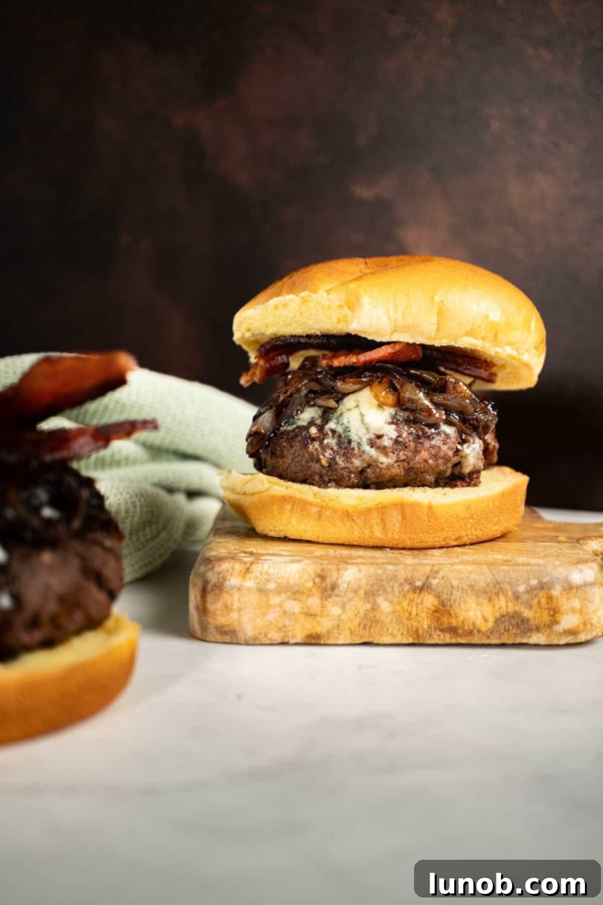 Bacon Blue Cheese Burger 2 A gourmet Bacon Blue Cheese Burger, topped with crispy bacon and sweet onion jam, served on a rustic cutting board, ready to be enjoyed.