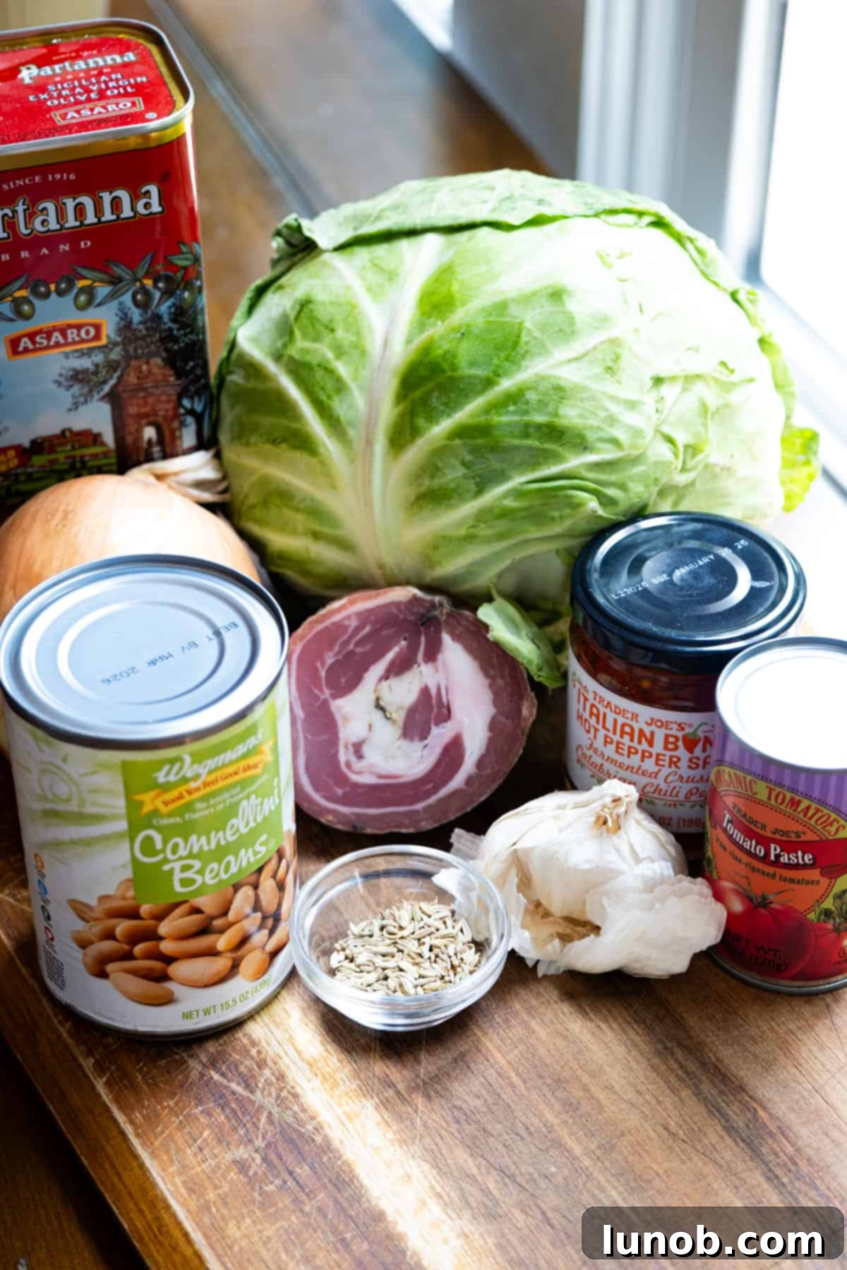Ingredients for Italian cabbage including pancetta, calabrian peppers, fennel seeds, tomato paste, cannellini beans, onion, and extra virgin olive oil laid out on a wooden board