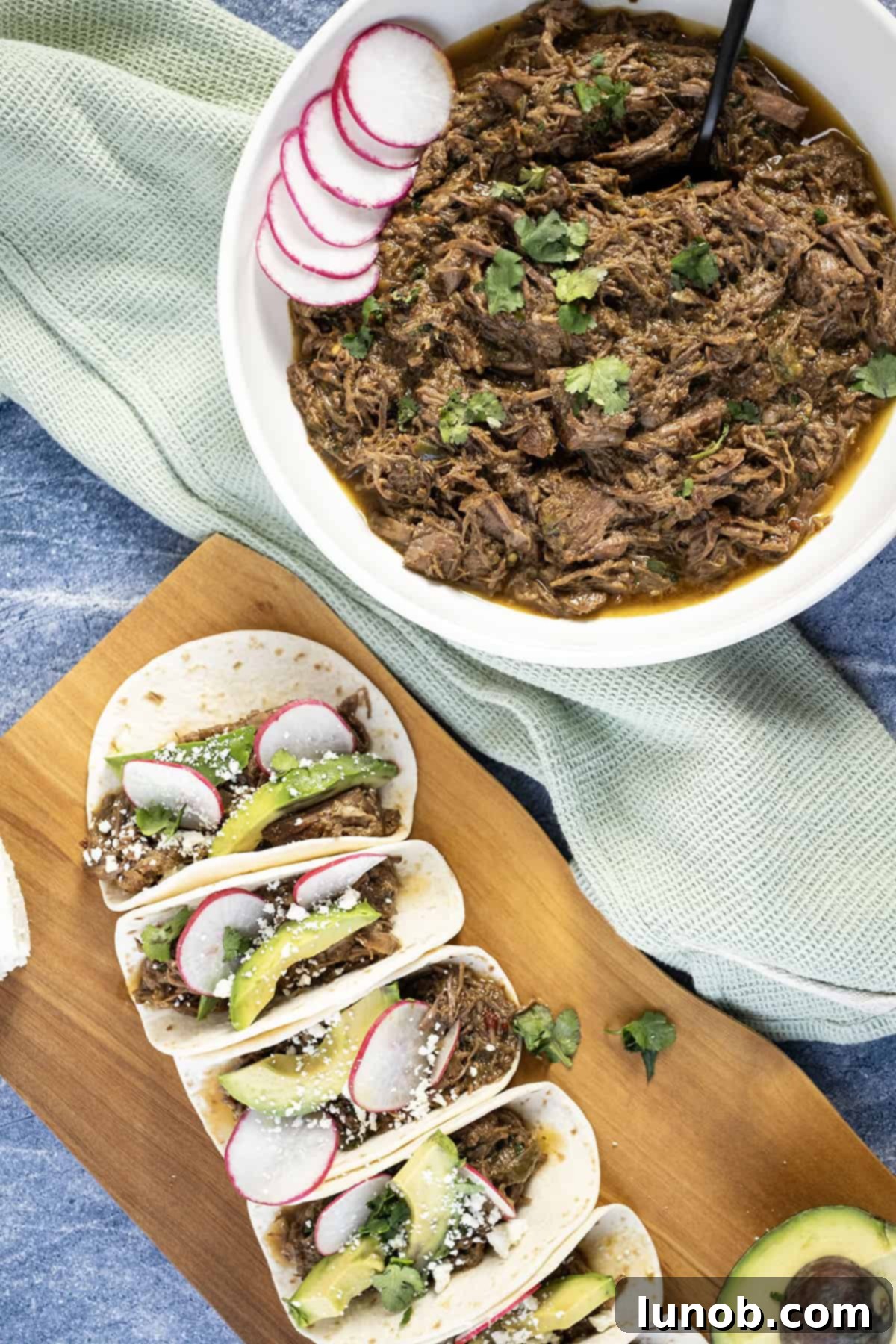 Beef barbacoa in a bowl alongside tacos with sliced radish, avocado, and cotija cheese. 