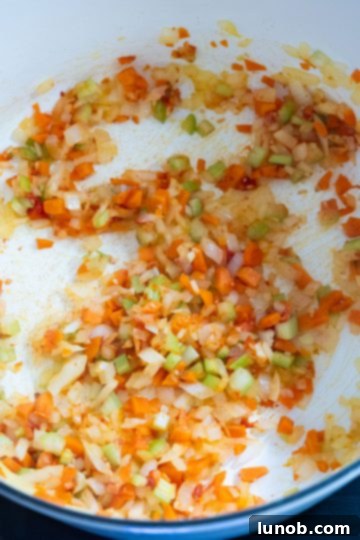 A close-up shot of chopped carrots, celery, and onion sautéing in a Dutch oven with tomato paste and minced garlic, forming a sofrito base.
