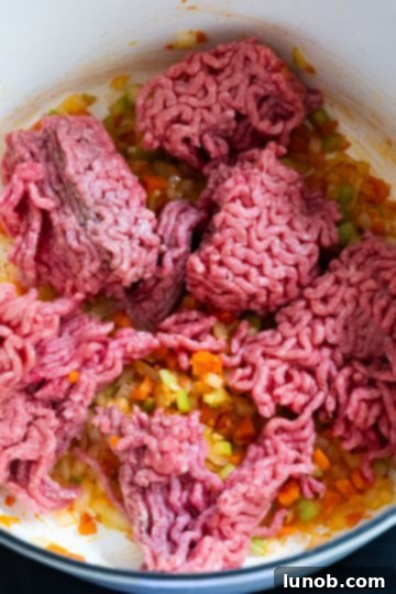 Ground beef being added to the rich sofrito base in a Dutch oven, ready to be browned for the stuffed pepper filling.