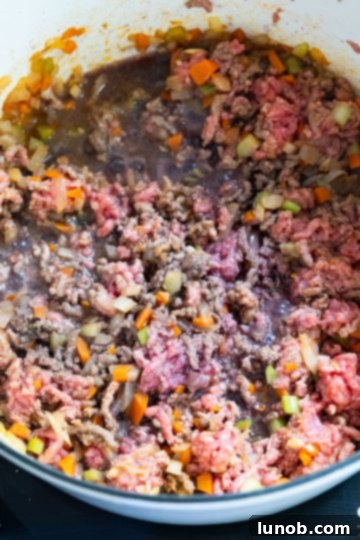 Partially browned ground beef simmering in red wine within a Dutch oven, creating a flavorful base for the tomato sauce.