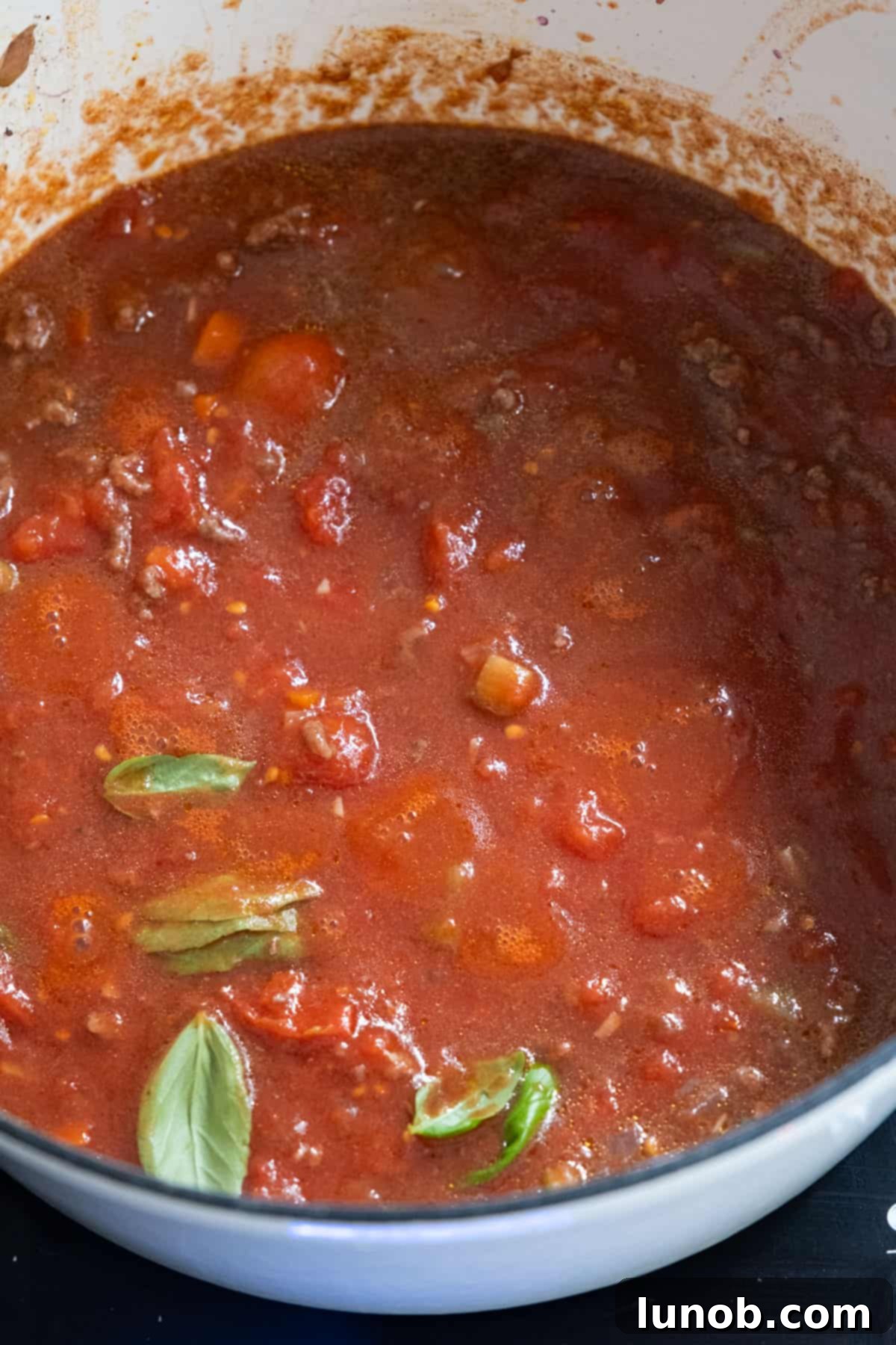 Crushed whole plum tomatoes being added to the simmering red wine and vegetable base in a Dutch oven, with the ground beef already removed.