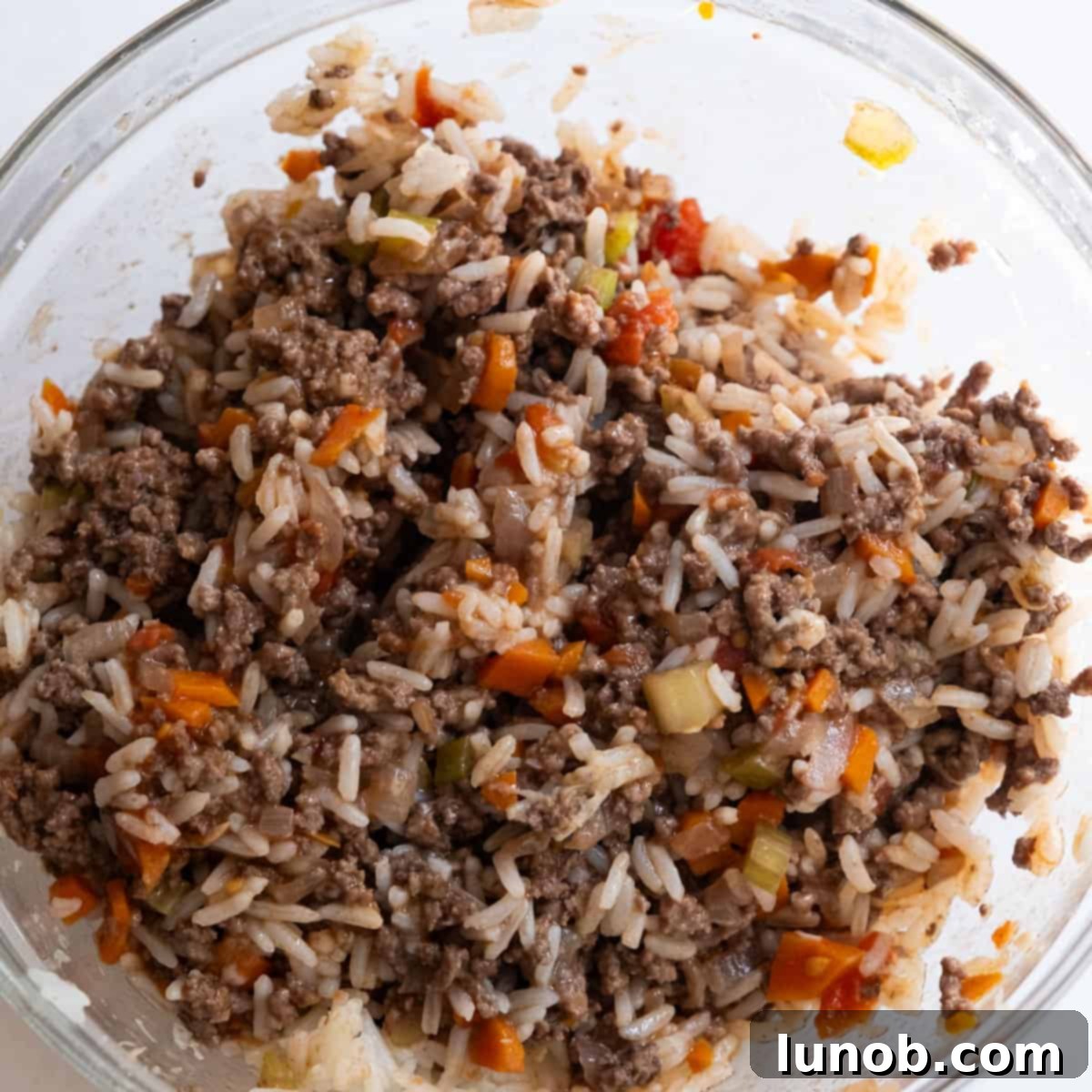 A bowl containing a mixture of ground beef, cooked rice, grated Parmesan cheese, and a spoonful of tomato sauce, ready to be combined for the stuffed pepper filling.