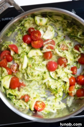 Freshly grated zucchini and halved cherry tomatoes being stirred into olive oil with fragrant minced garlic in a pan.
