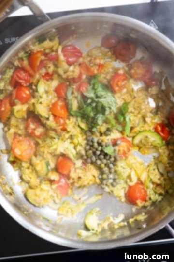 Fresh capers and torn basil leaves being folded into the simmering zucchini and cherry tomato sauce.