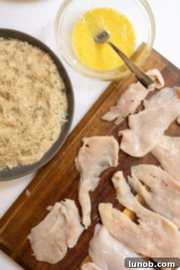 Thinly sliced chicken breast laid out on cutting board with bowl of whisked eggs and seasoned breadcrumbs.