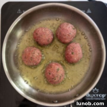 Raw meatballs gently placed into a hot saucepan containing olive oil, beginning the frying process.