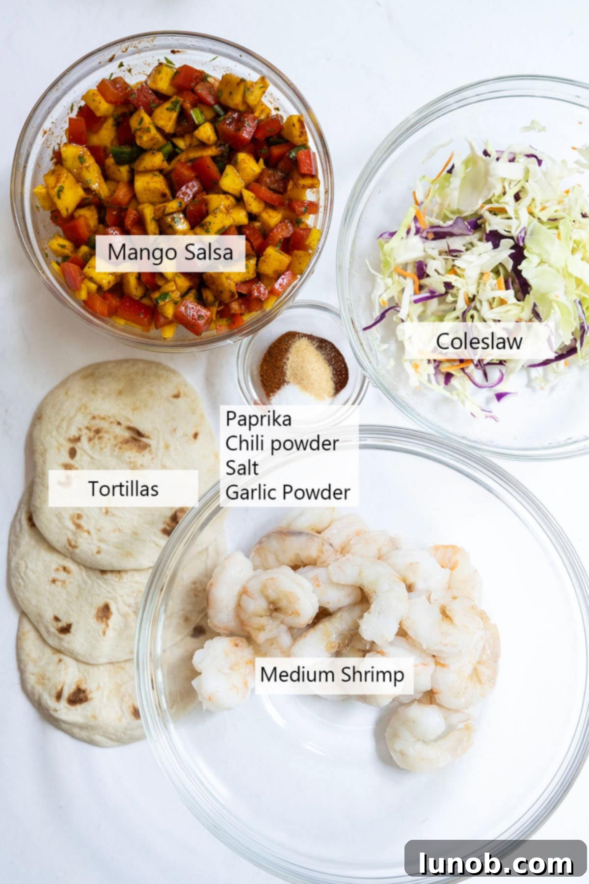 Ingredients for shrimp tacos, including mangoes, red pepper, jalapeno, cilantro, lime, and shrimp, all laid out on a wooden board.