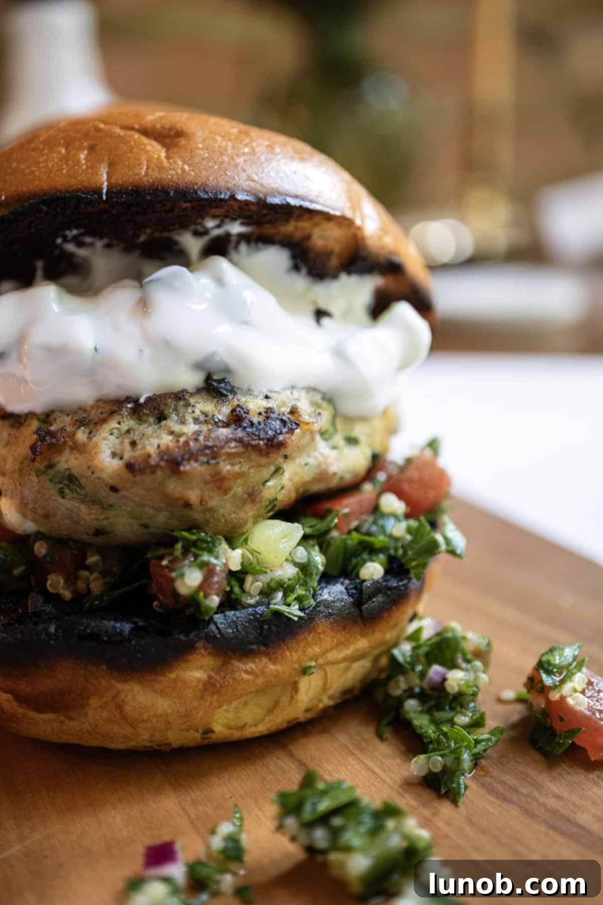 Close-up of a perfectly grilled Greek chicken burger on a toasted bun, generously topped with fresh tabbouleh and drizzled with creamy tzatziki.