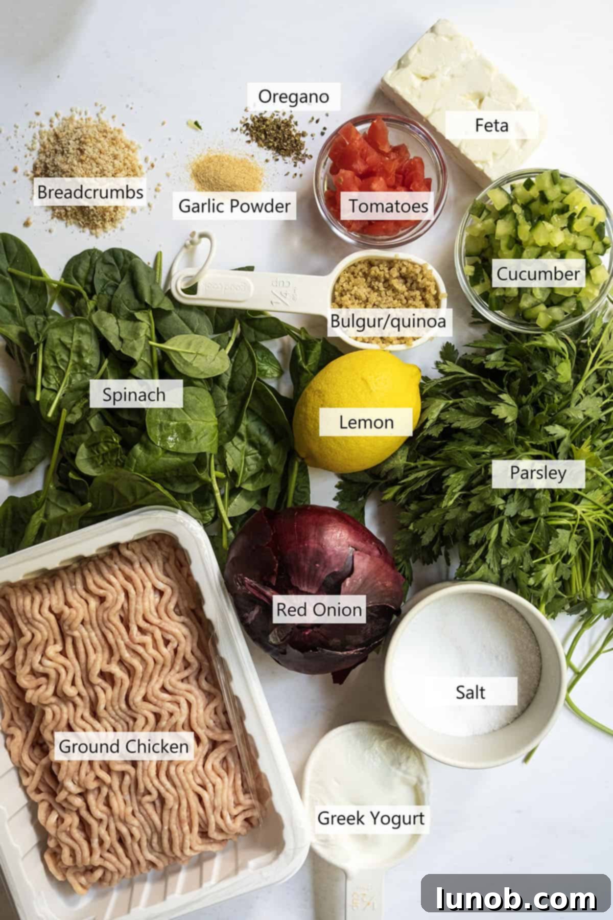 Fresh ingredients laid out for Greek chicken burgers, including ground chicken, feta, spinach, oregano, and components for tabbouleh and tzatziki.