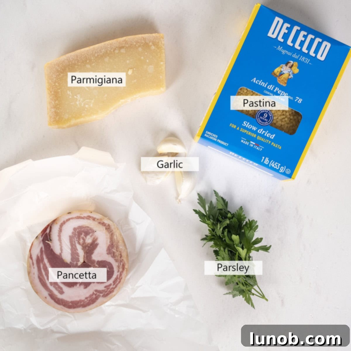Fresh ingredients laid out on a kitchen counter for Creamy Pastina Carbonara, including uncooked pastina, slices of pancetta, garlic cloves, and a block of Parmigiana cheese.