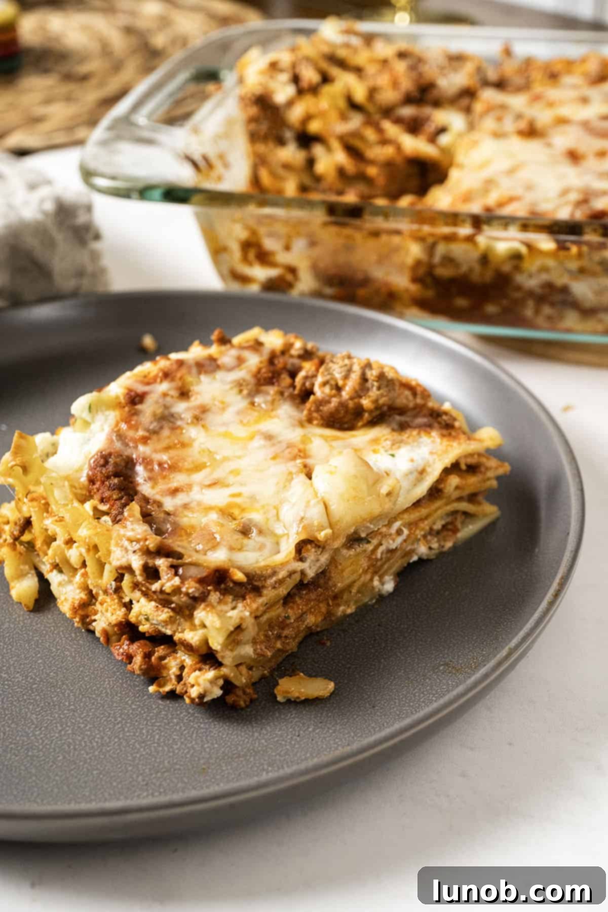 A generous square of authentic Italian lasagna on a white plate, with a baking dish filled with more lasagna in the background.