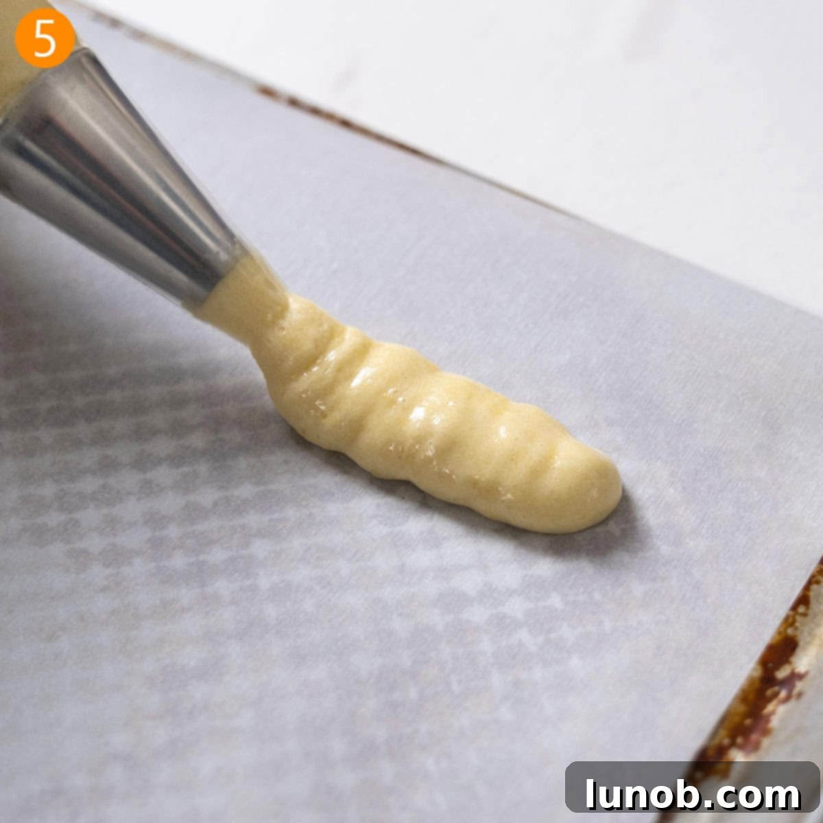 Ladyfinger batter being piped in uniform 4-inch strips onto parchment paper on a baking sheet.