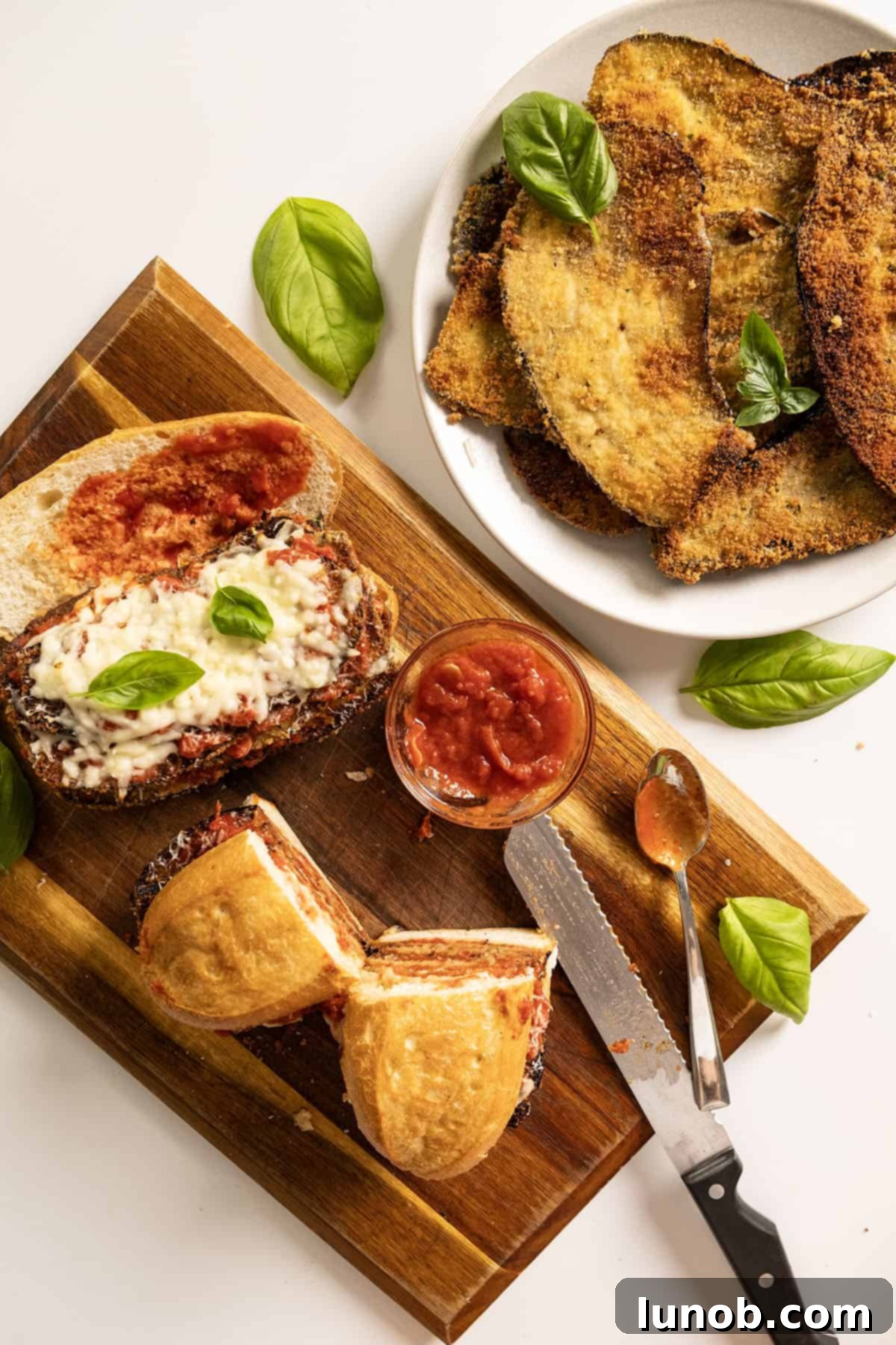 Two eggplant parm sandwiches on a cutting board with a plate of fried eggplant.