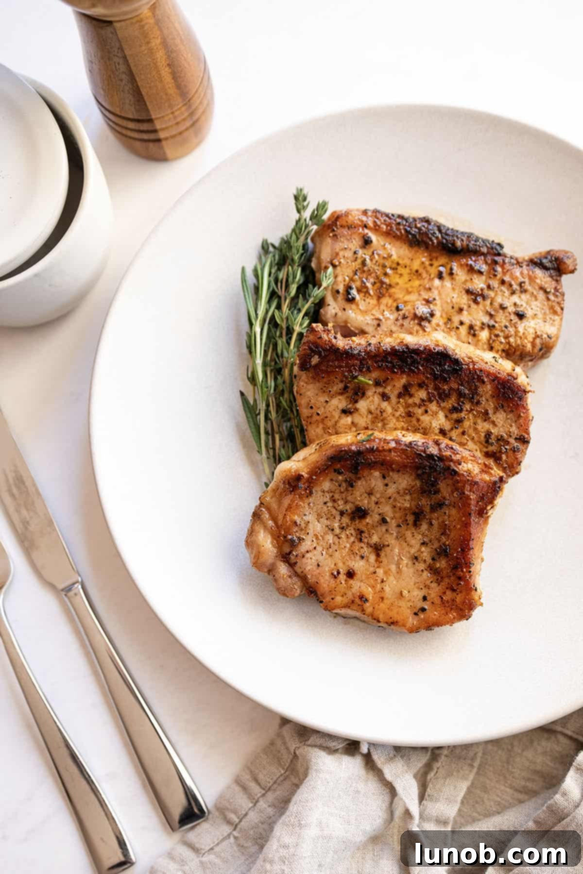 Ultimate Cast Iron Boneless Pork Chops 2 Two perfectly cooked boneless pork chops resting on a white plate, garnished with fresh rosemary and thyme sprigs.