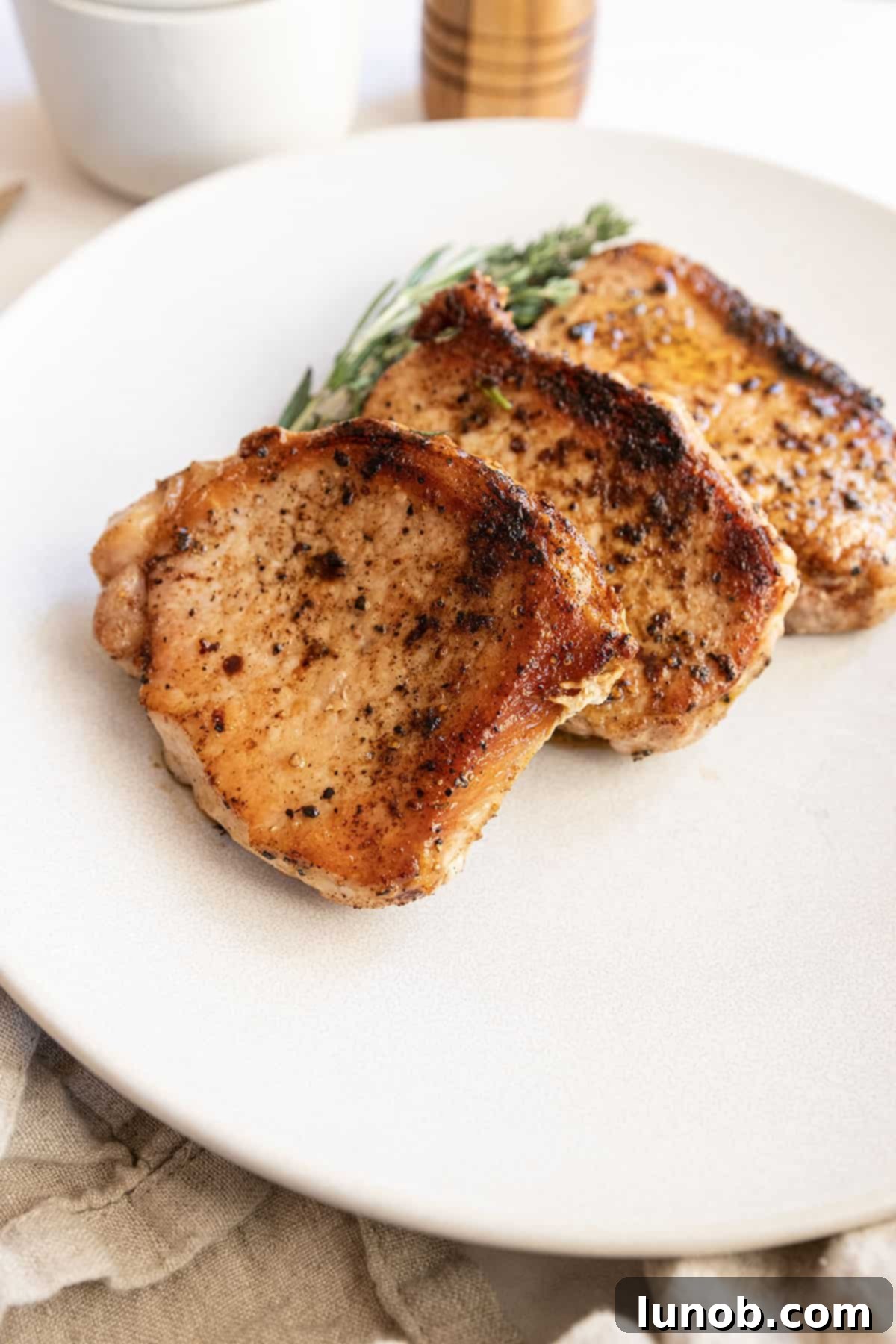 Ultimate Cast Iron Boneless Pork Chops 9 Two boneless pork chops, beautifully seared and basted, garnished with fresh rosemary and thyme on a white plate.