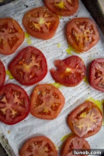 Caprese Eggplant Melt 4 Thickly sliced tomatoes on a parchment-lined baking sheet, seasoned with olive oil, salt, pepper, and garlic powder, ready for roasting.