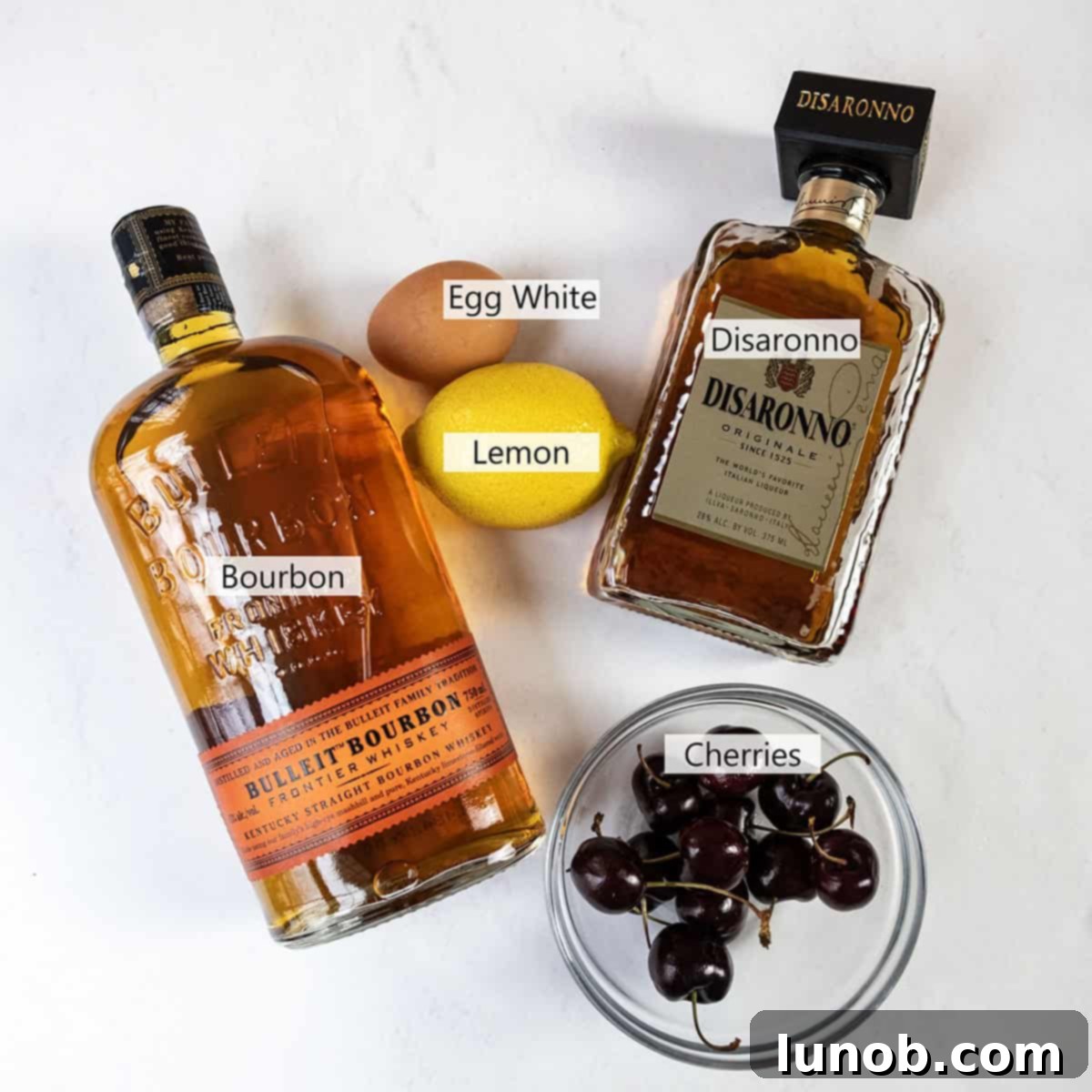 Fresh ingredients laid out for making a Cherry Amaretto Sour cocktail: Amaretto, Bourbon, fresh cherries, lemon, and an egg.