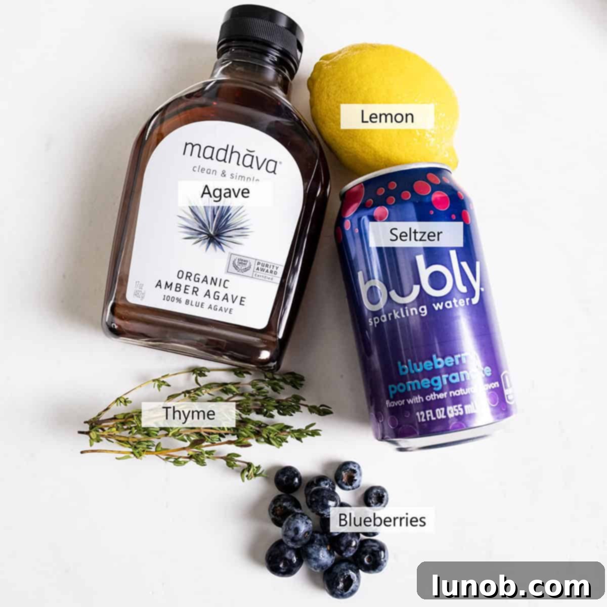 Blueberry Blitz Mocktail 3 Fresh ingredients laid out for making the blueberry mocktail: seltzer, agave syrup, blueberries, a lemon, and a sprig of thyme.