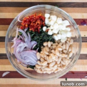 A large bowl filled with colorful ingredients: chopped kale, cannellini beans, mozzarella pearls, sundried tomatoes, and thinly sliced red onion.