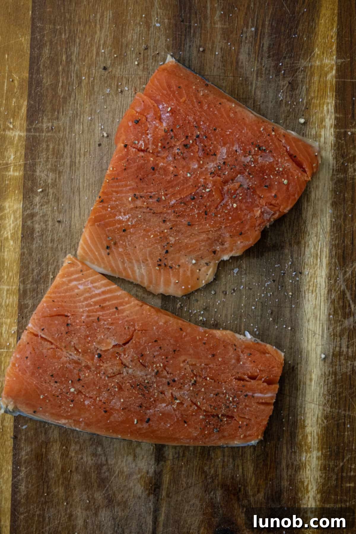 Two salmon fillets being seasoned generously with salt and pepper on a cutting board.