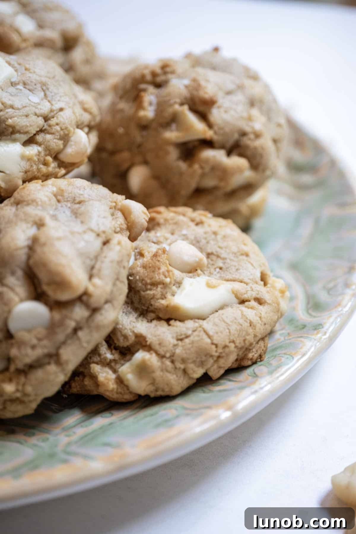 Soft Baked White Chocolate Macadamia Delights 11 A stack of golden-brown white chocolate macadamia nut cookies on a white plate, ready to be eaten.