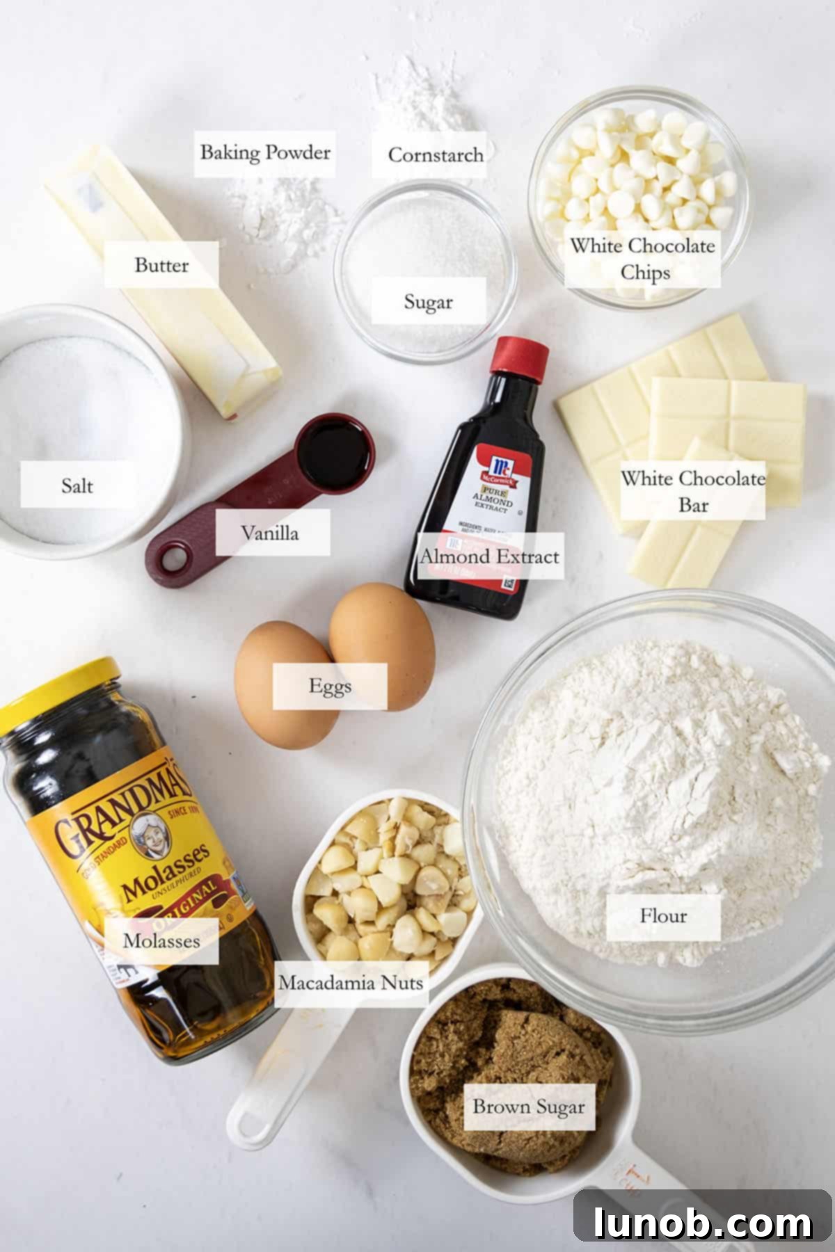 Soft Baked White Chocolate Macadamia Delights 3 A collection of ingredients for white chocolate macadamia nut cookies, including flour, butter, sugars, eggs, macadamia nuts, and white chocolate.