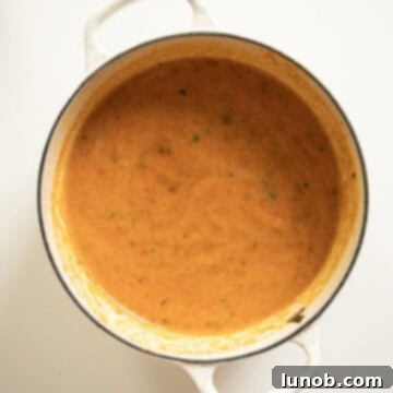 Rich Roasted Garlic and Tomato Cream Soup 9 A smooth, vibrant red tomato soup is being blended in a large pot using an immersion blender, indicating the final stage of achieving its creamy texture.