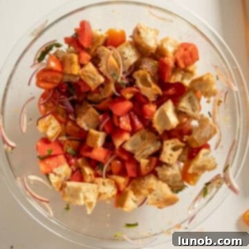 Cubed bread soaking in a bowl with mixed tomatoes, red onion, and basil, absorbing the flavorful juices.
