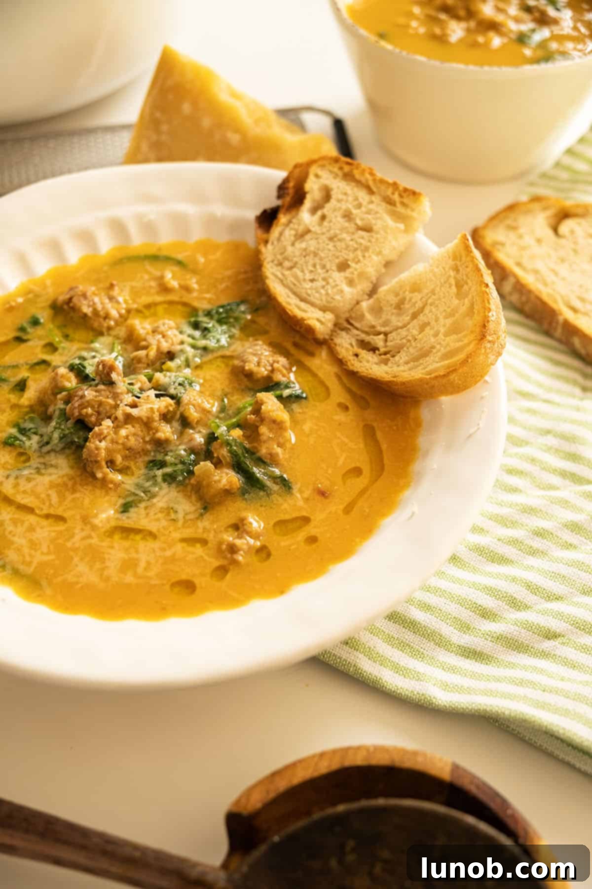 A steaming bowl of creamy butternut squash soup with crumbled Italian sausage, garnished with fresh herbs, served alongside crusty Italian bread on a rustic wooden surface.