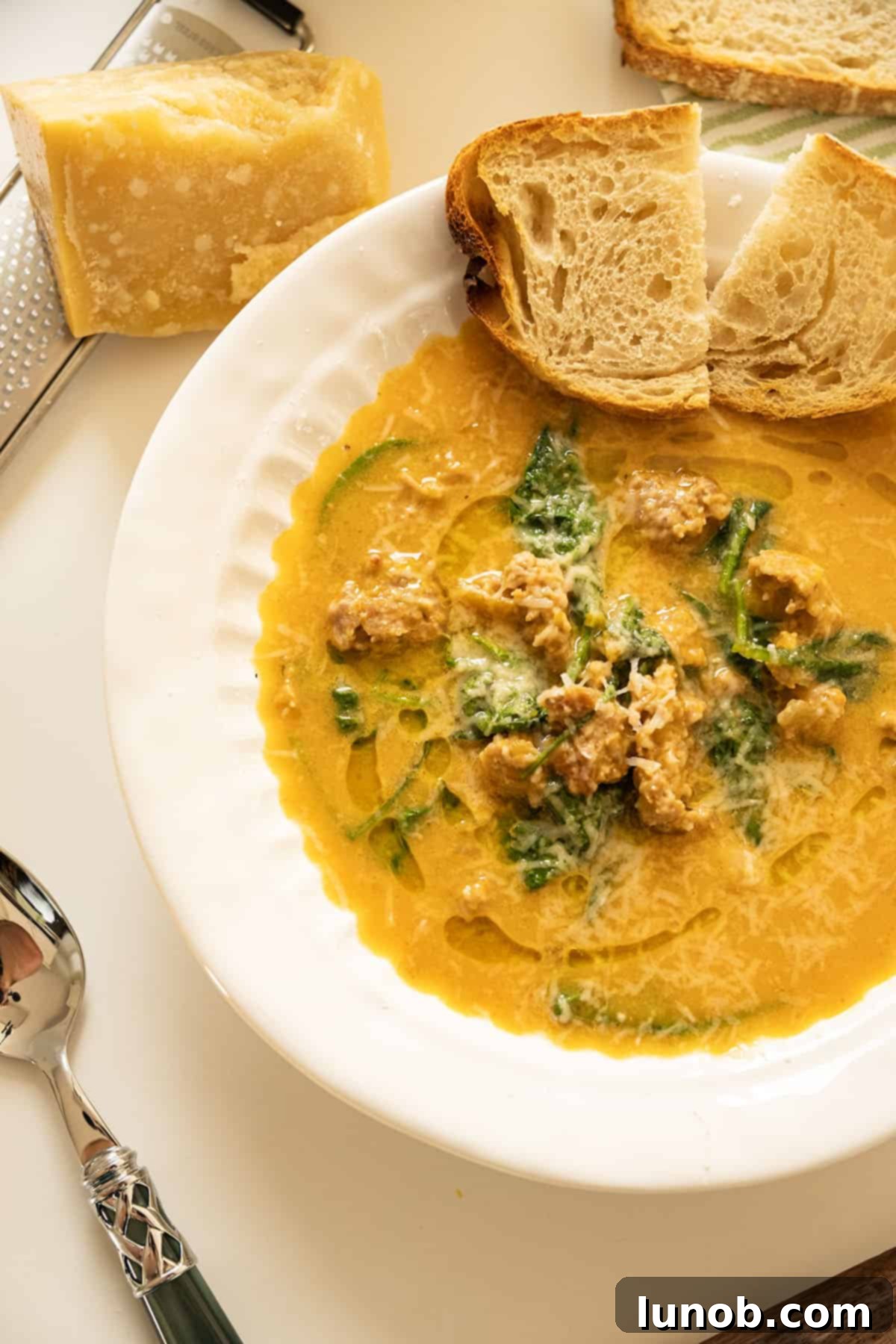 A bowl of creamy butternut squash soup with sausage, accompanied by a basket of crusty Italian bread and a small wooden spoon.